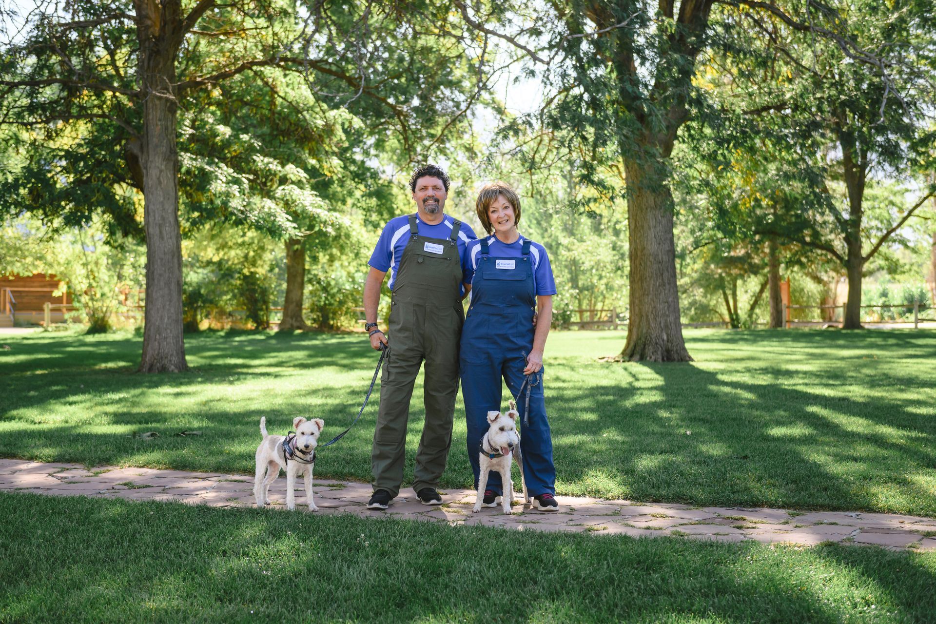 A pair in matching blue shirts and overalls stands on a stone path in a green park, each holding a small dog on a leash.