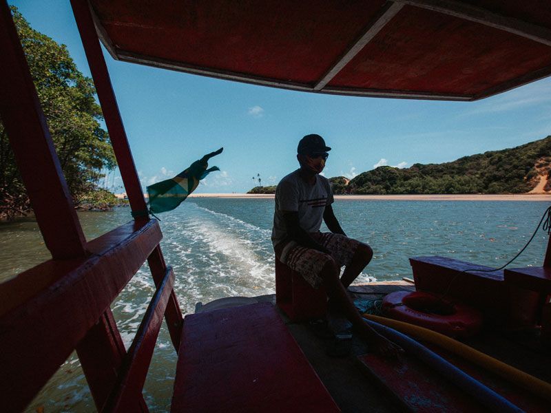 Homem em um barco vermelho navegando em direção à praia; céu azul, bandeira e árvores verdes.