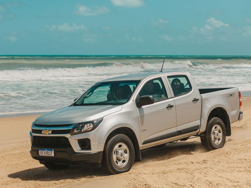Caminhonete Chevrolet S10 prateada em uma praia de areia, com o oceano ao fundo.