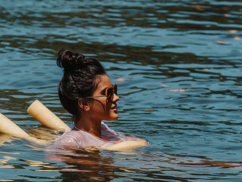 Mulher de óculos escuros flutua na água, cabelo preso em coque, segurando um macarrão de piscina.