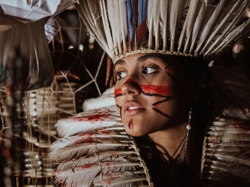 Pessoa com pintura facial e cocar de penas olha para o lado. Veste uma roupa ornamentada com penas, em um ambiente com pouca luz.