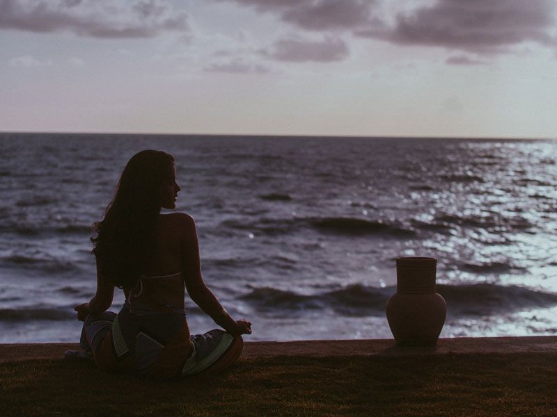 Mulher meditando em posição de lótus, de frente para o oceano ao pôr do sol. Um jarro de barro está próximo.