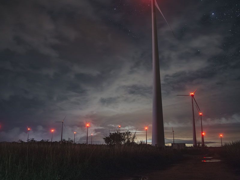 Turbinas eólicas à noite com luzes vermelhas, sob um céu nublado e estrelado.