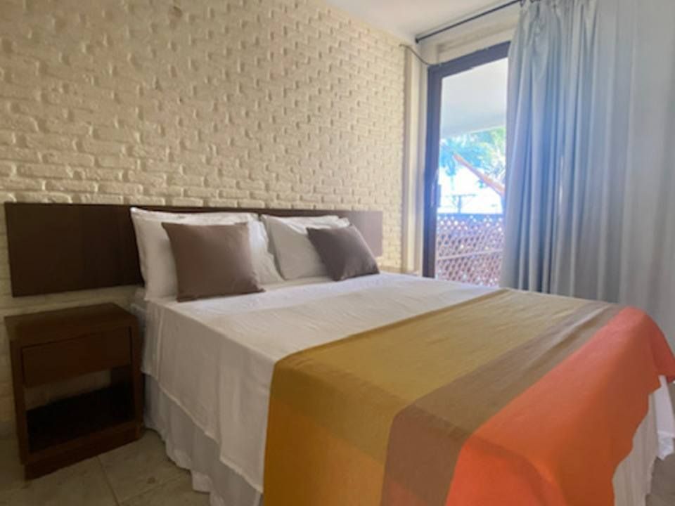 Quarto com parede de tijolos brancos, cama de casal, travesseiros marrons, mesa de cabeceira e acesso à varanda.