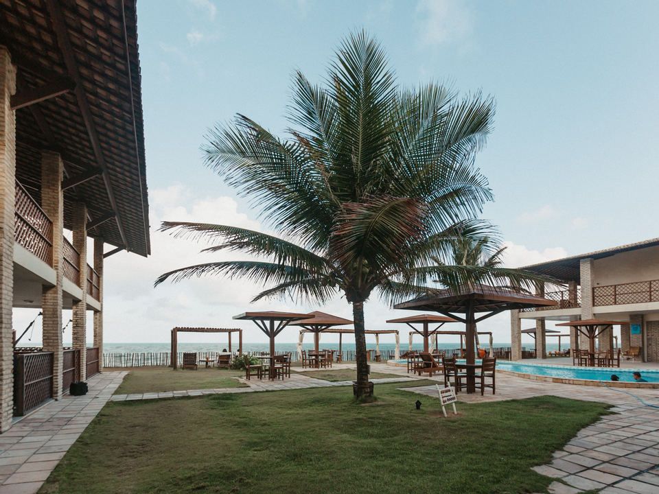 Resort de praia com palmeiras, gramado, gazebos de madeira, piscina e vista para o mar.