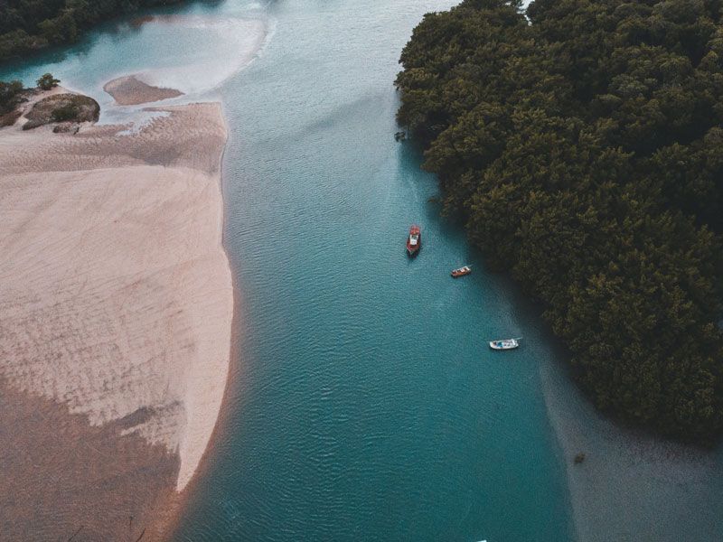 O rio serpenteia por uma paisagem costeira com barcos, praia de areia e árvores verdejantes. Água turquesa.