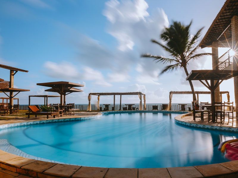 Piscina em um resort com água azul, pérgulas de madeira, palmeiras e vista para o mar em um dia ensolarado.
