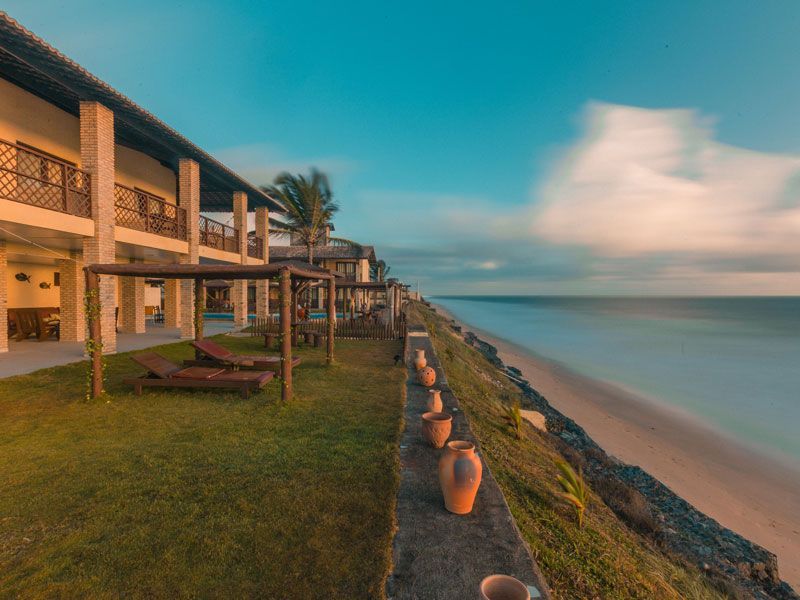 Resort costeiro com vista para o mar. Longa exposição da praia, gramado e edifício com pérgulas de madeira. Tons de azul e dourado.