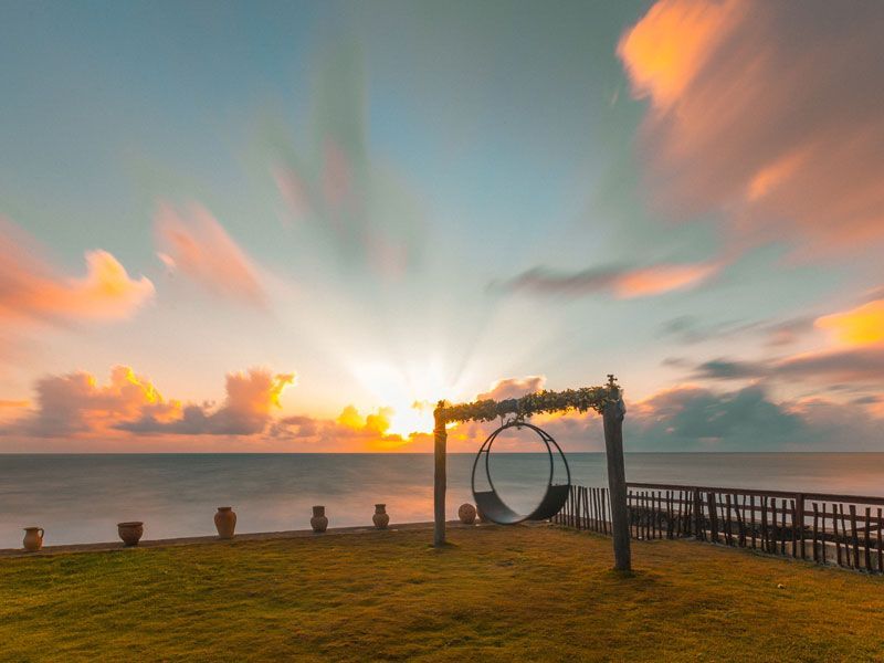 Pôr do sol sobre o oceano com balanço, grama, vasos e nuvens; cores laranja e azul.