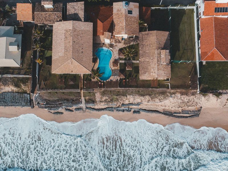 Vista aérea de casas à beira-mar com piscina, areia e ondas do oceano.