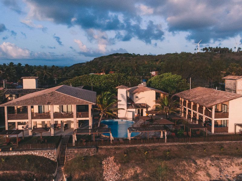 Resort tropical com edifícios, piscina e vegetação exuberante, visto de cima sob um céu nublado.