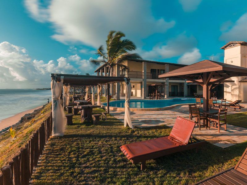 Villa de luxo à beira-mar com piscina, espreguiçadeiras e pérgulas sob um céu azul nublado.