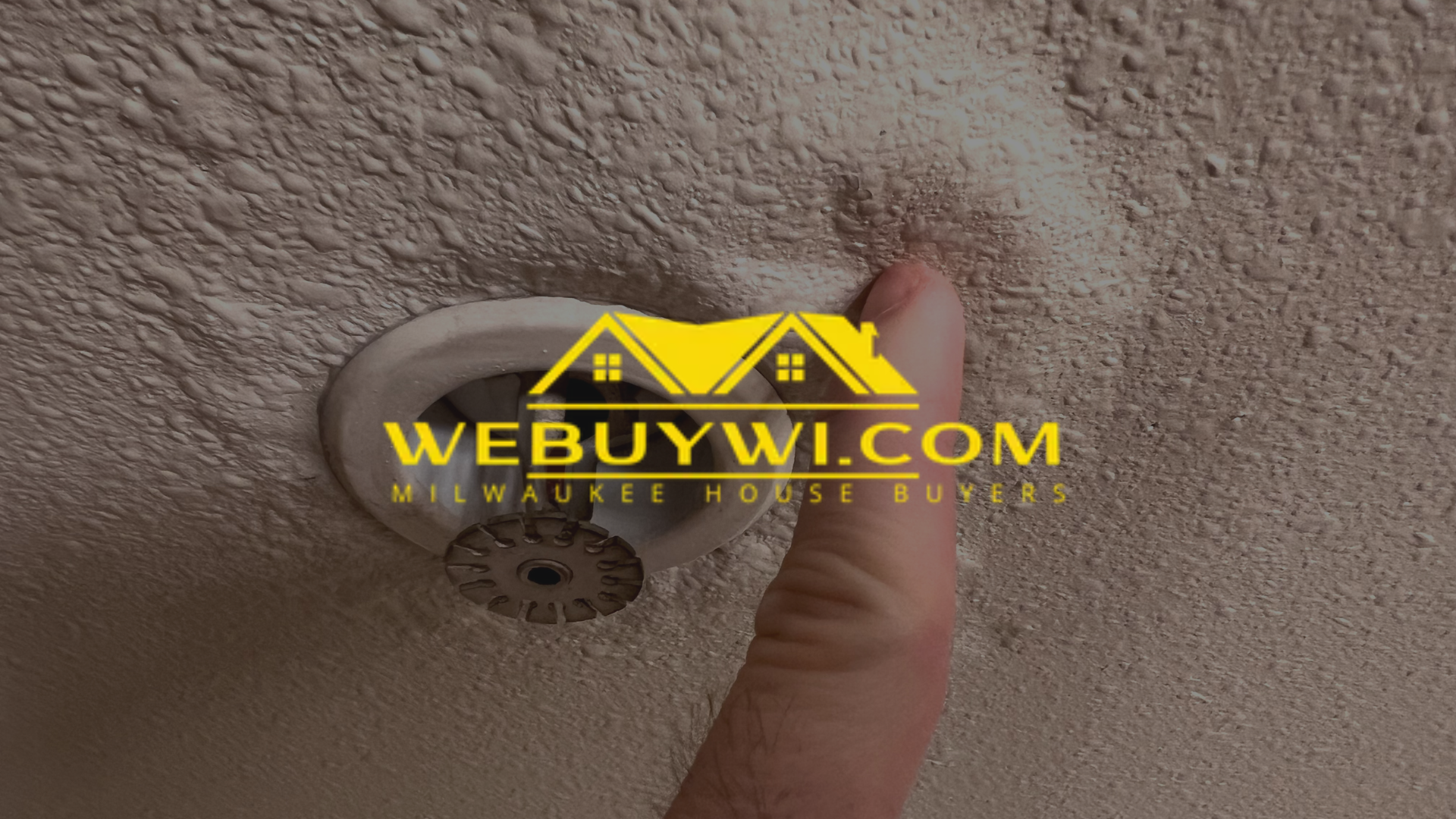 Hand touching a discolored ceiling with a sprinkler system, likely water damage.