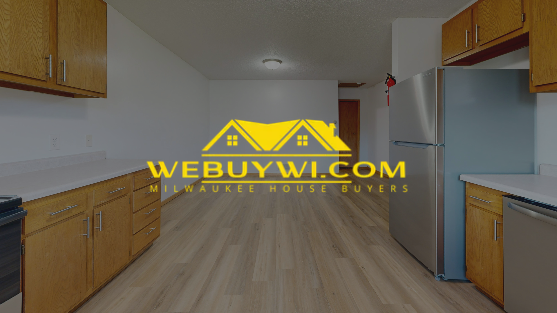 Kitchen interior with wooden cabinets, light countertops, and stainless steel refrigerator. Logo overlay: WeBuyWi.com.