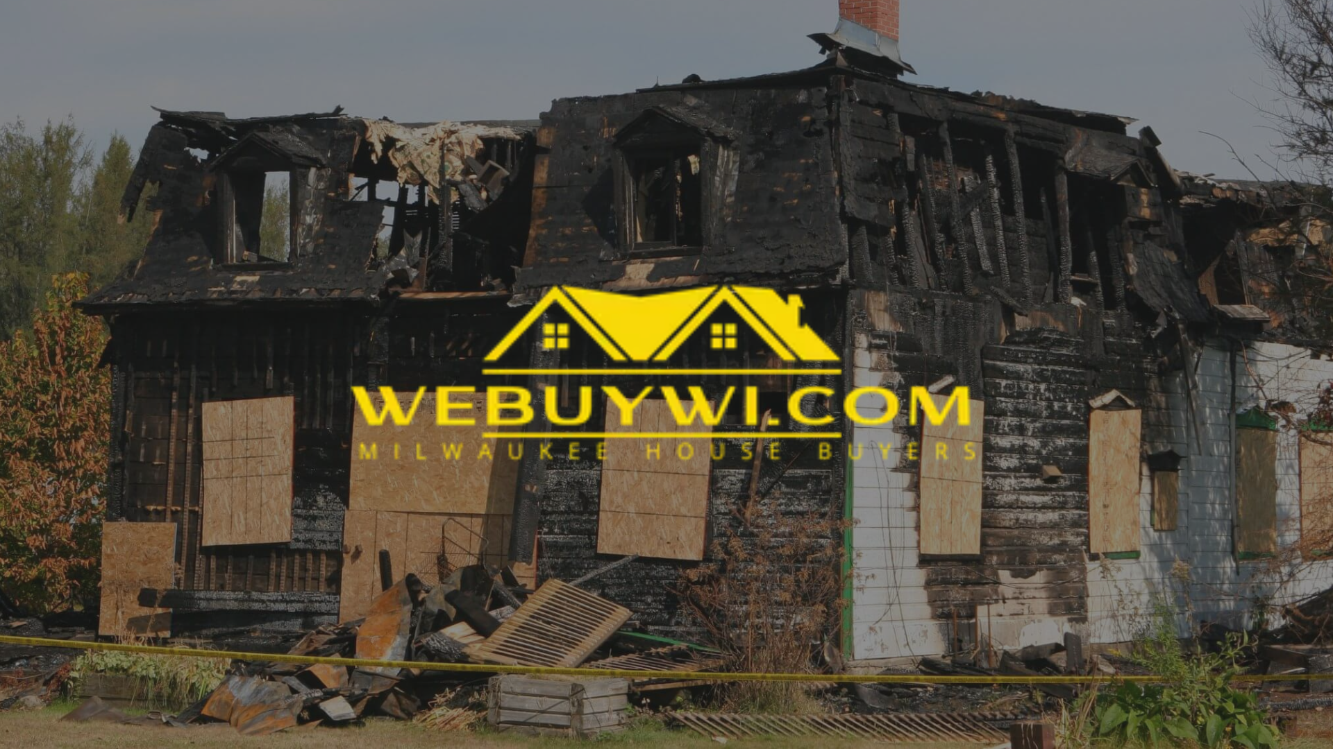A severely burned house with boarded-up windows, likely after a fire, features the