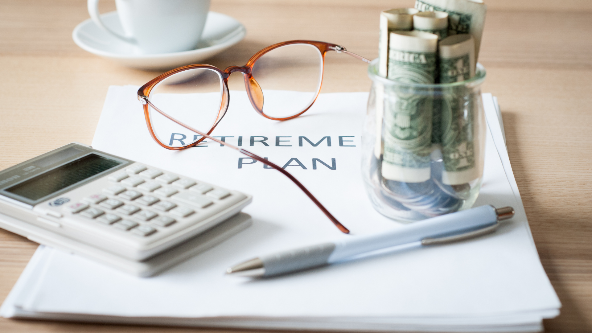 A calculator , pen , glasses , and a glass of money are on a table.