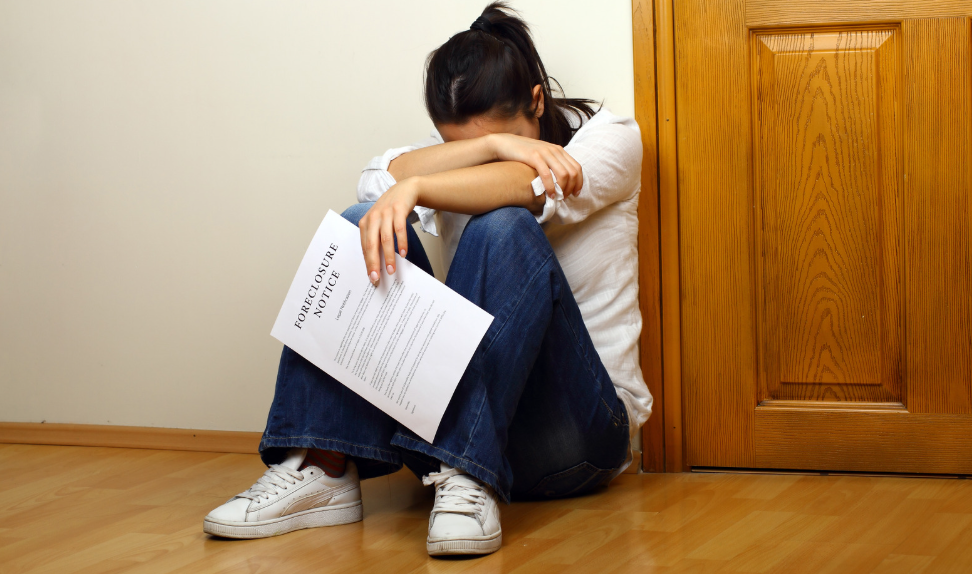 A woman is sitting on the floor with her head down holding a foreclosure notice