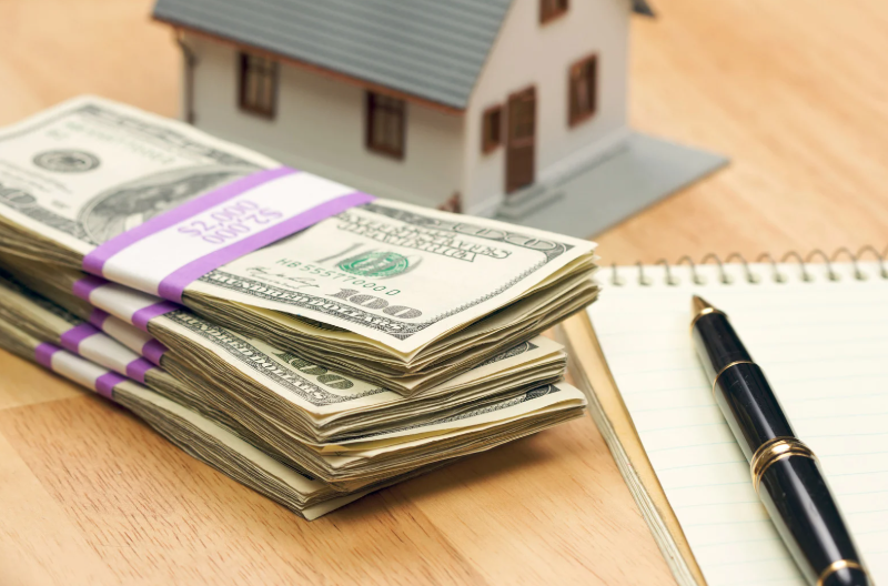 Stacks of cash, toy house, notepad, and pen on a wooden surface.