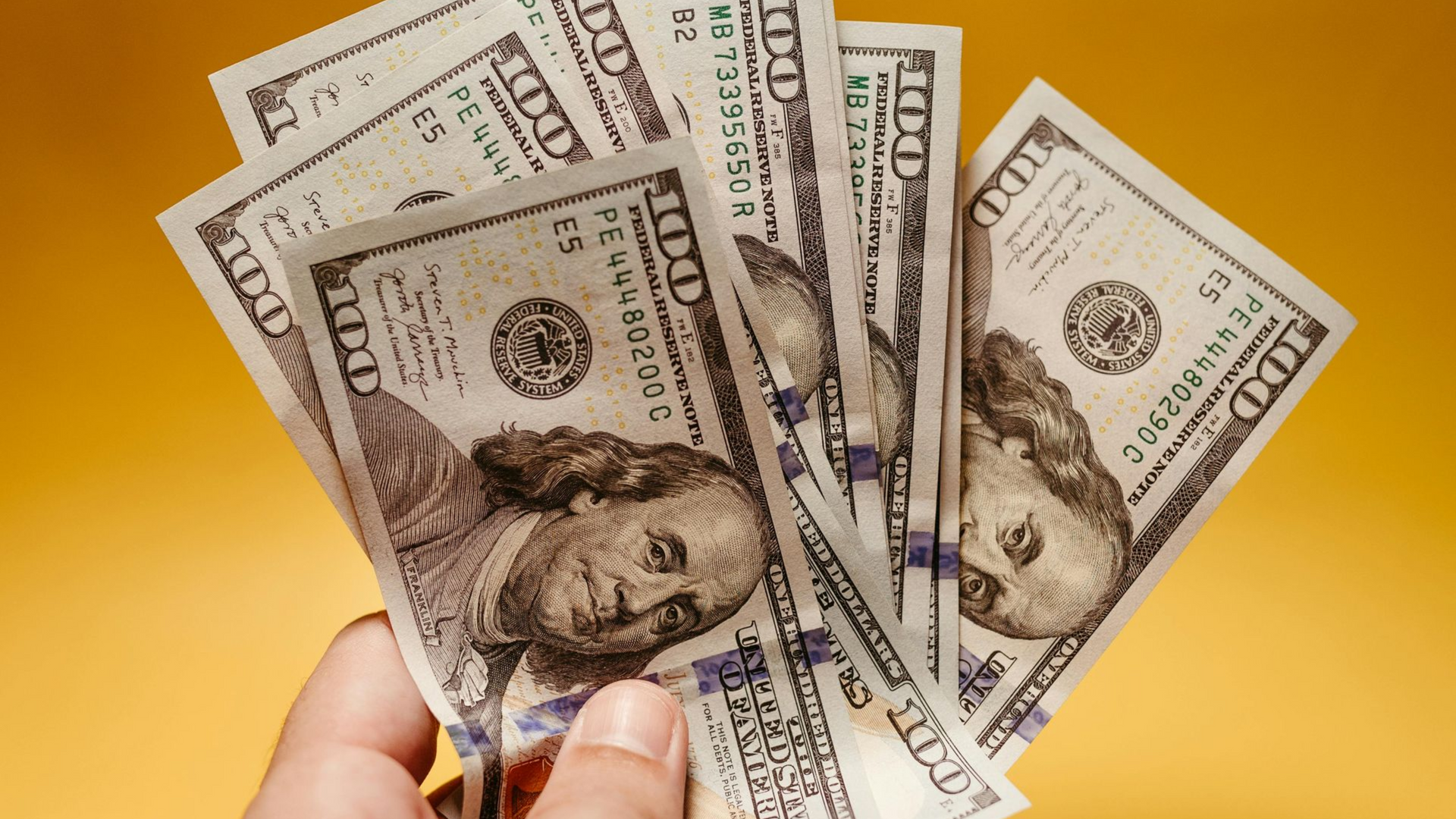 Hands fanning several U.S. dollar bills against a bright yellow background