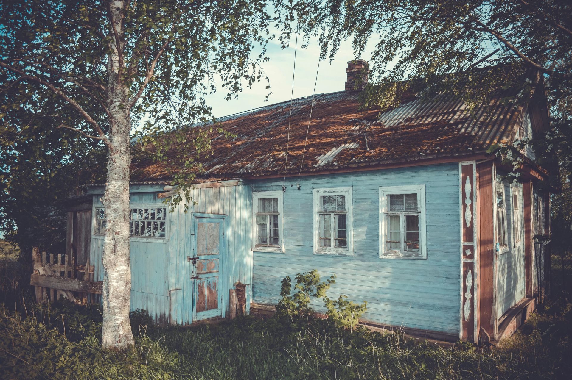 An aged, light blue wooden house with a weathered tile roof stands among trees in a grassy, sunlit yard.