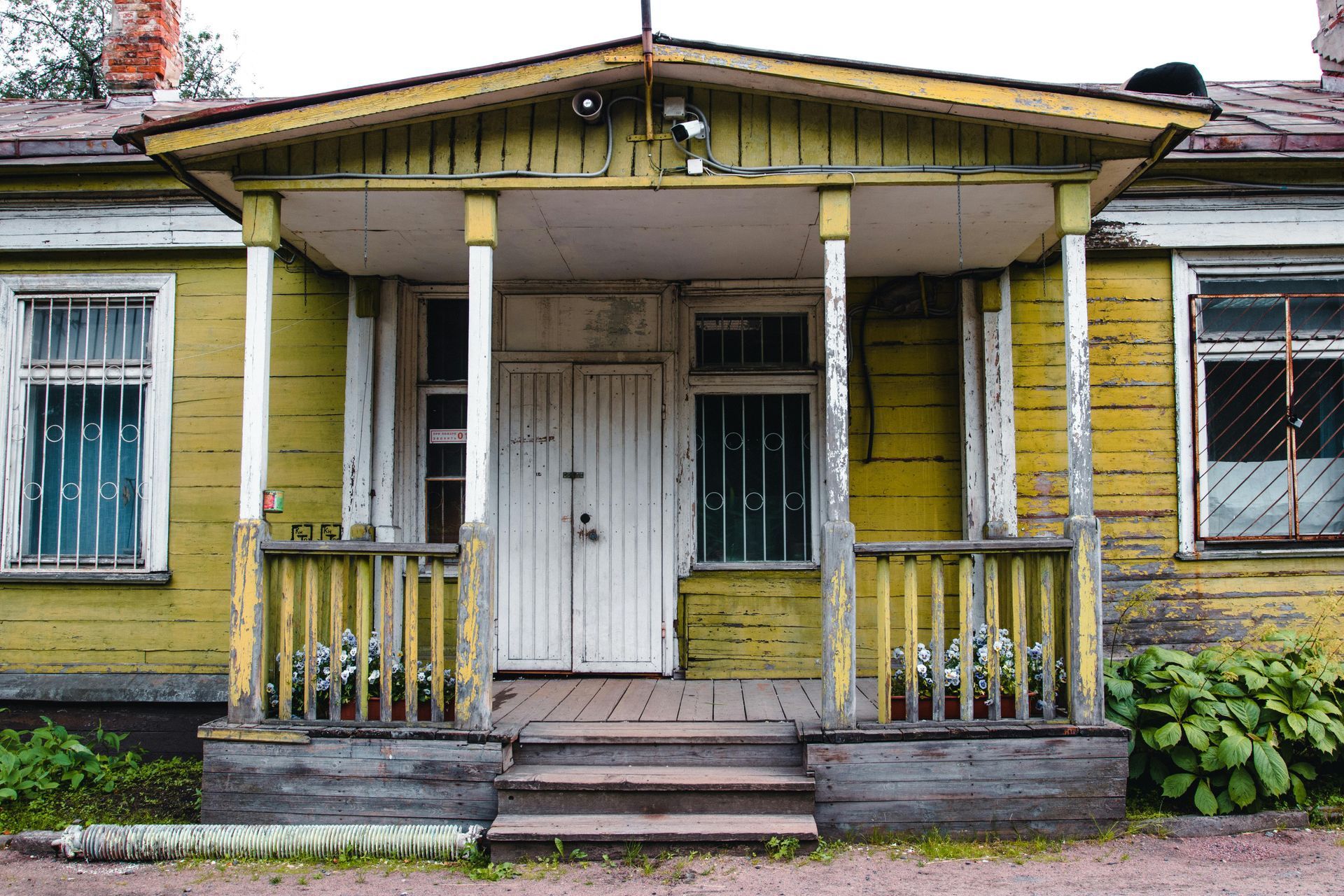 A weathered yellow wooden house with a white double-door entrance, a covered porch with stairs, and barred windows.