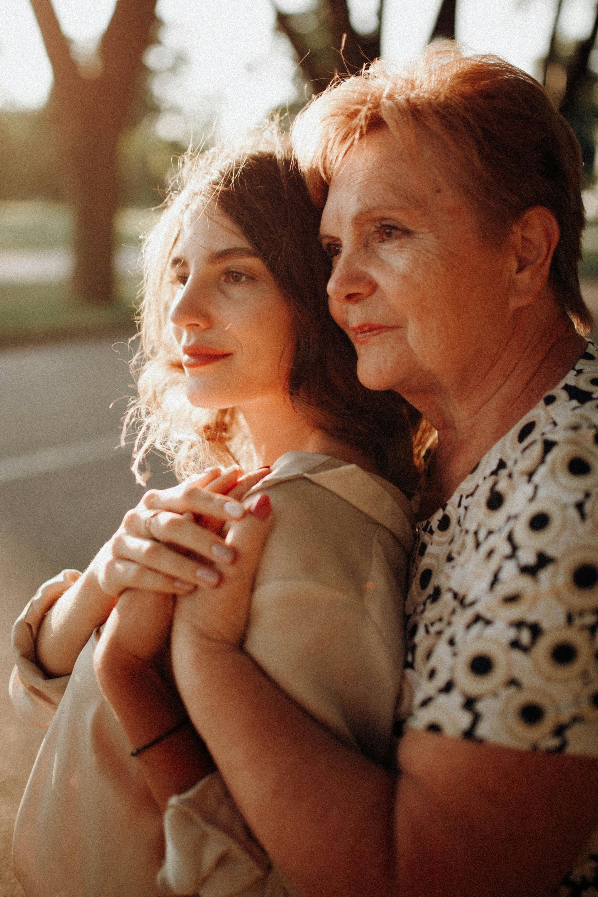 Two women sitting close together in warm sunlight, gently holding hands and looking ahead.