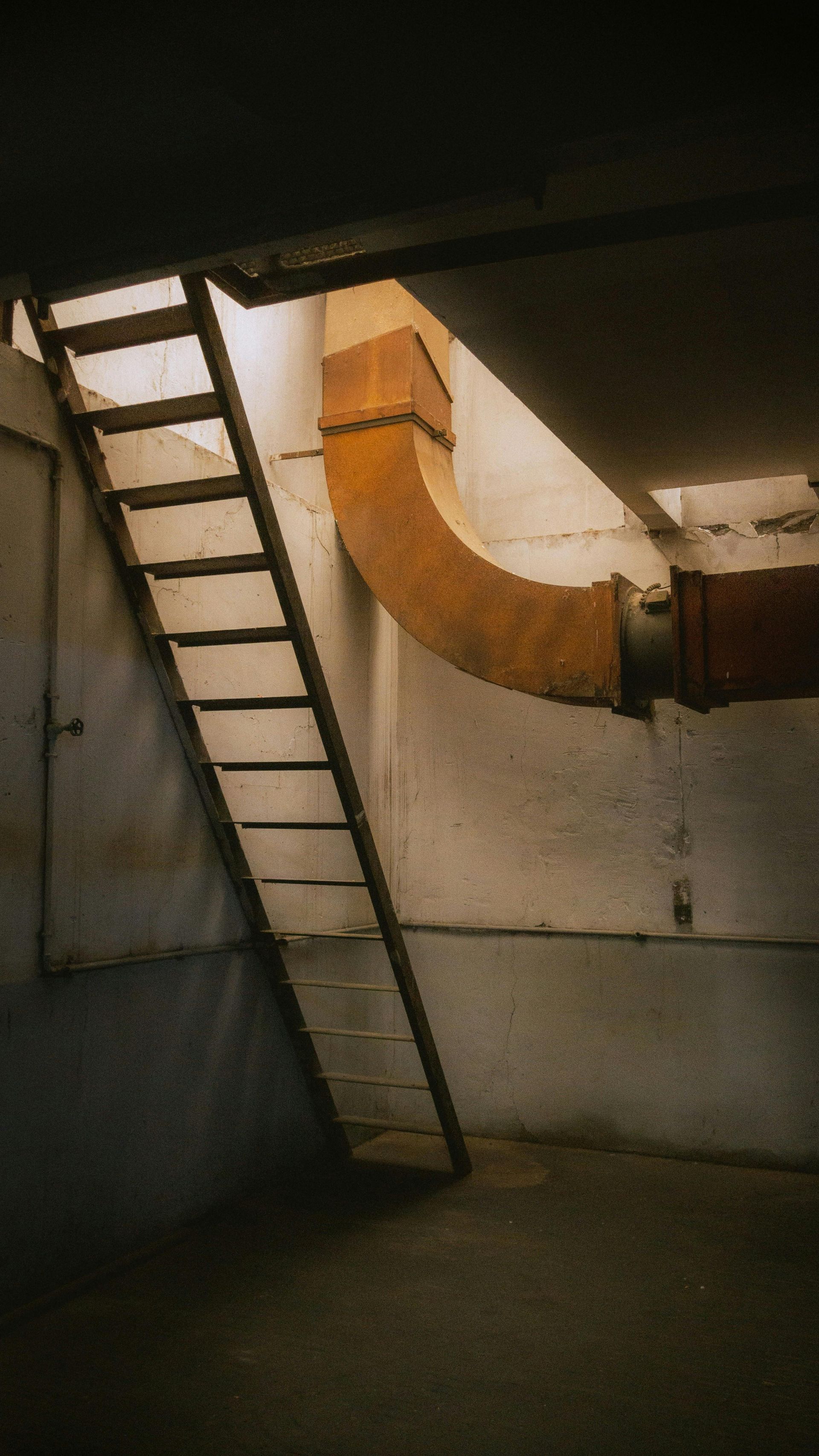 Dim basement with a metal ladder and large orange ventilation duct on the wall