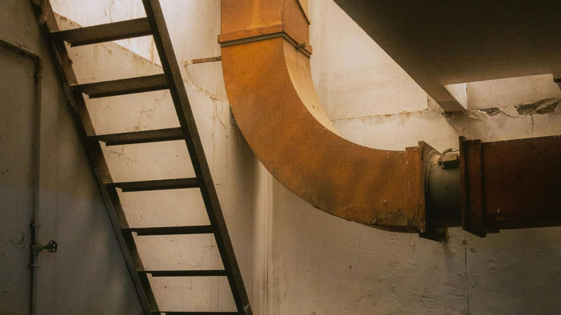 Industrial wall with a black ladder beside a large curved orange pipe and rusted ductwork