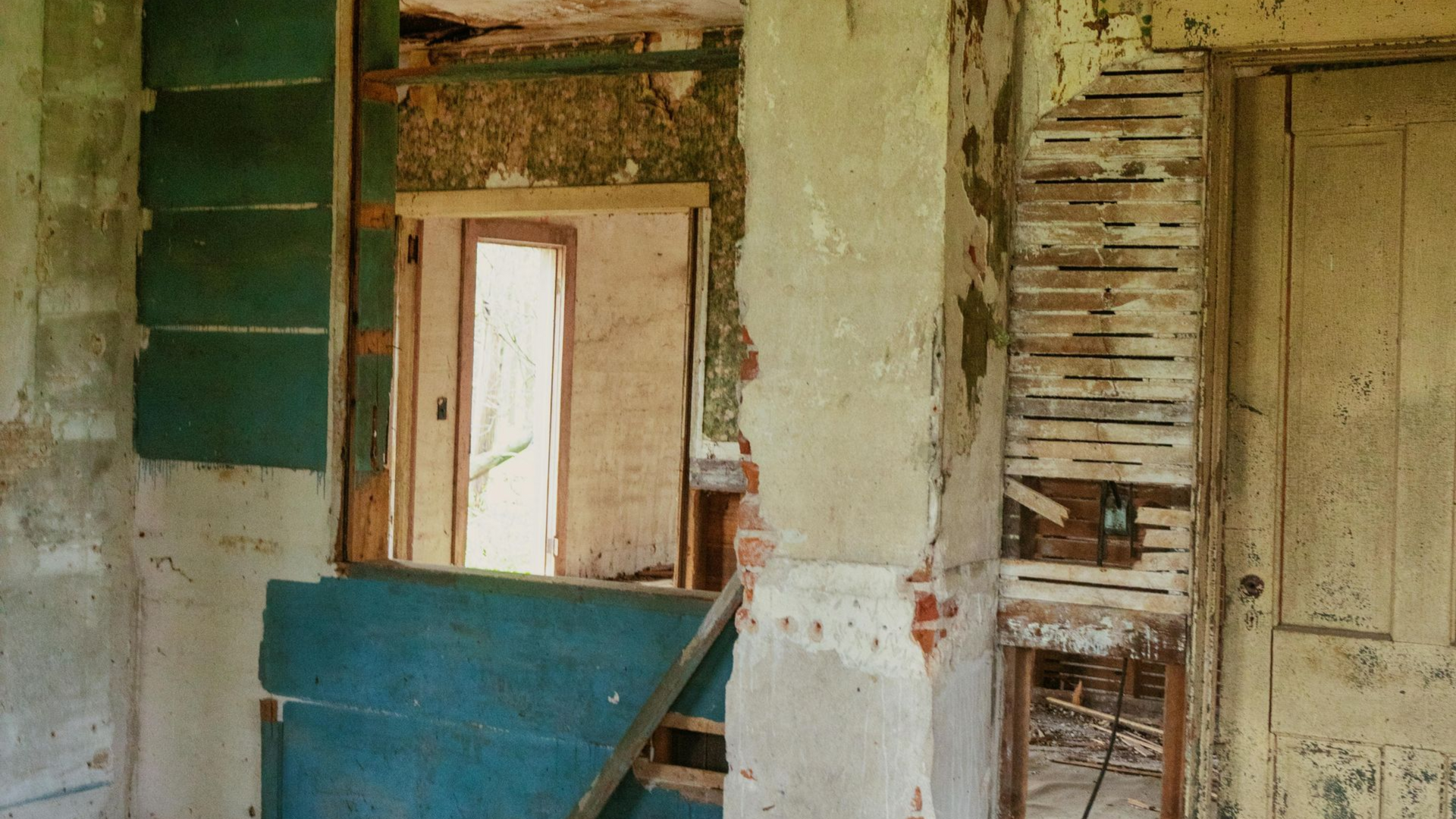 Interior of a dilapidated room featuring exposed lath, green painted panels, and a view into an adjacent empty space.
