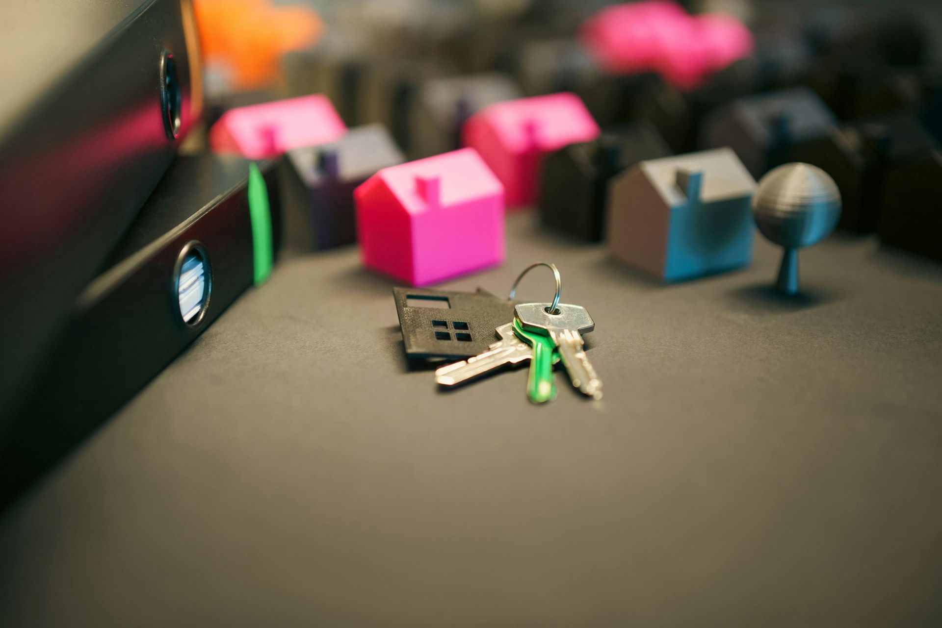 A set of keys on a ring with a small house charm, resting near miniature 3D-printed houses and black binders.