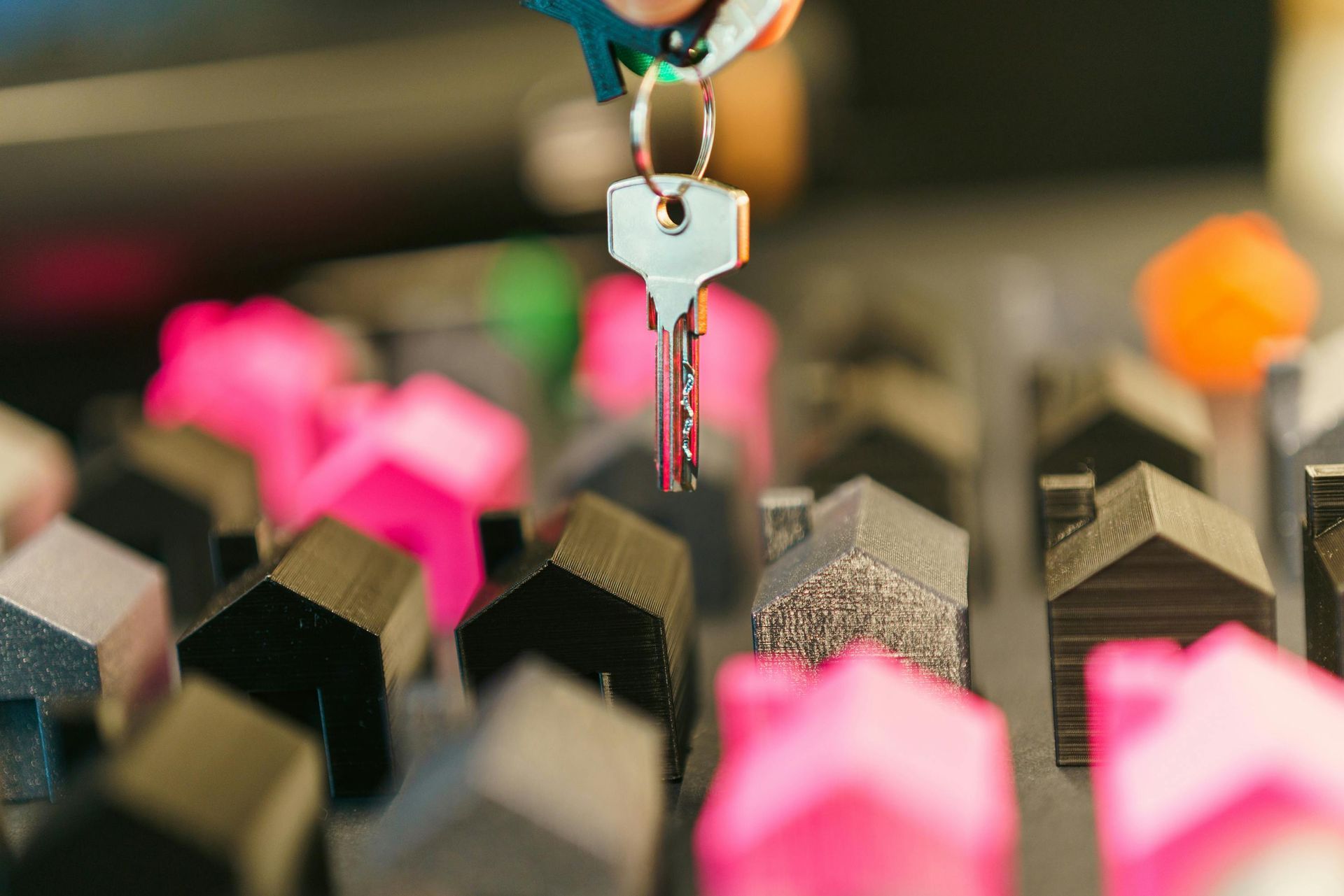 A hand holds a metal key over a scattered collection of small, colorful house models in pink, black, and orange.