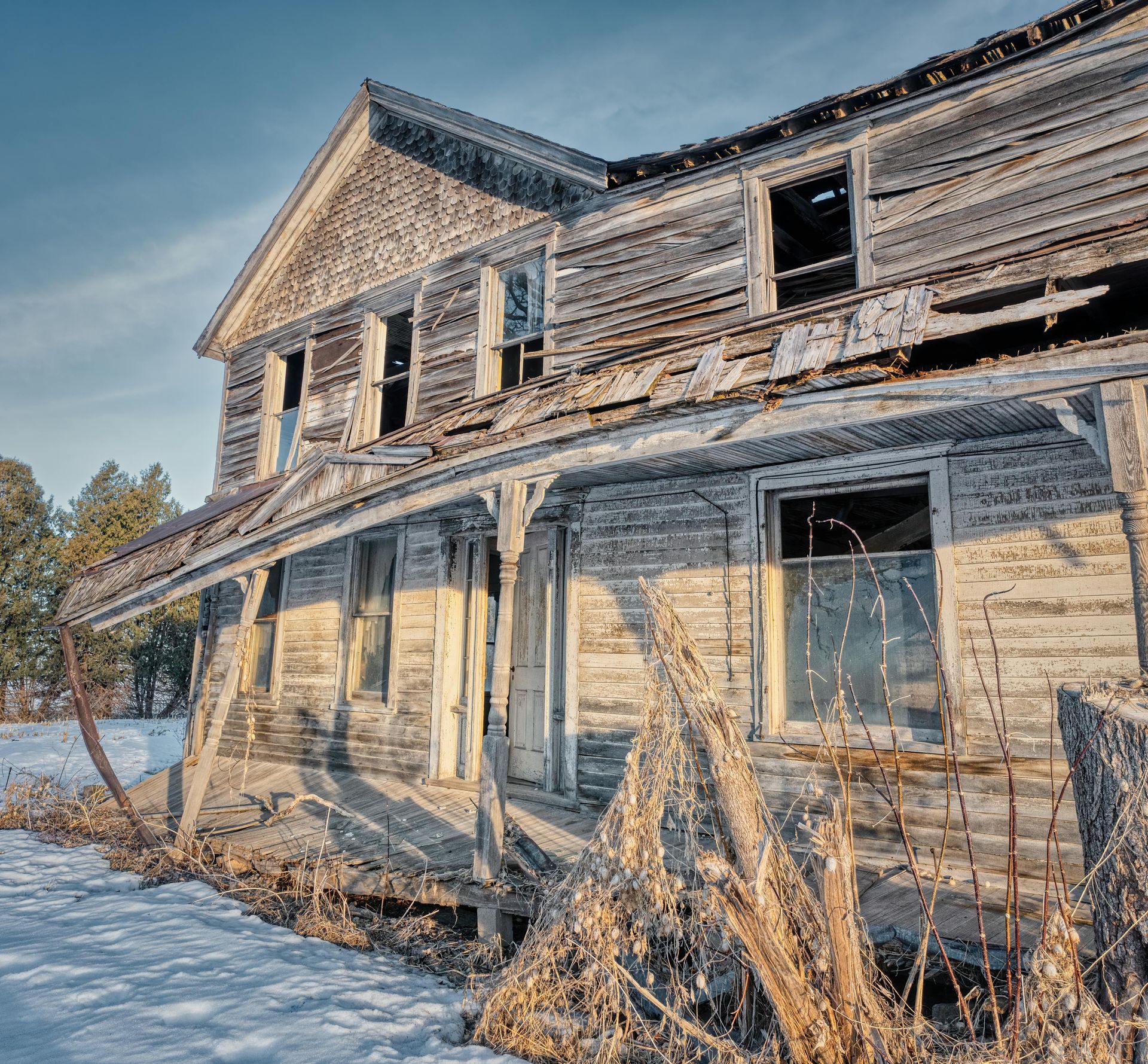 Weathered two-story farmhouse with peeling white paint and broken windows in a snowy rural setting