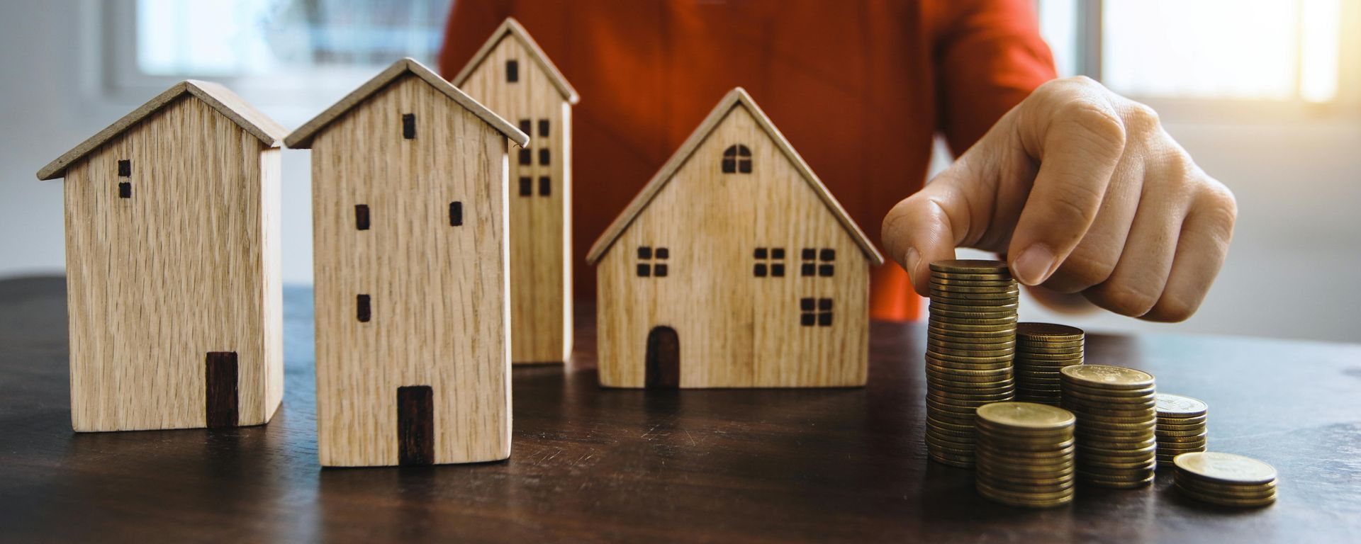 Person stacking coins near wooden house models, symbolizing real estate investment.