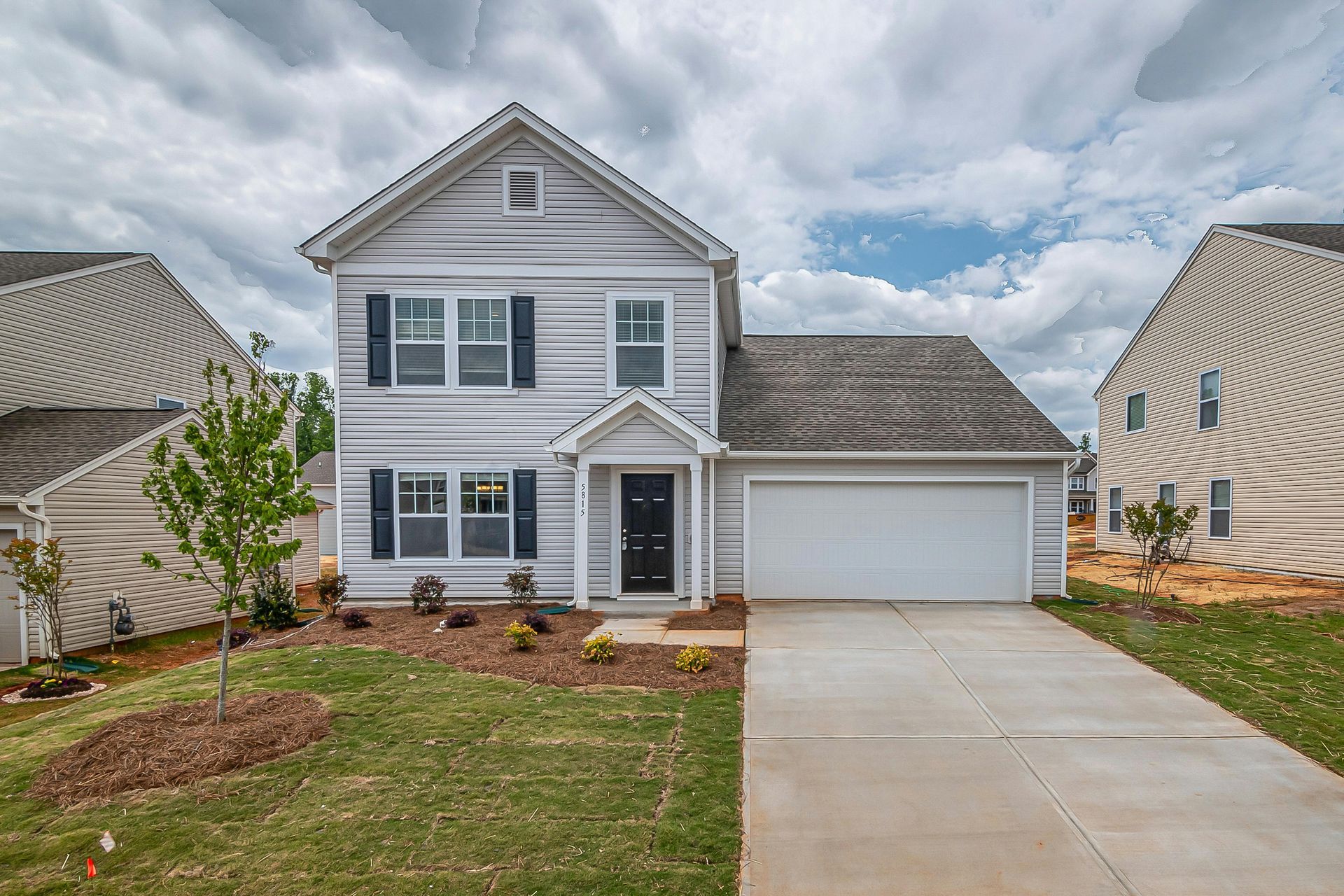 A two-story, light-grey suburban house with a white garage door, black shutters, and a front lawn under a cloudy sky.