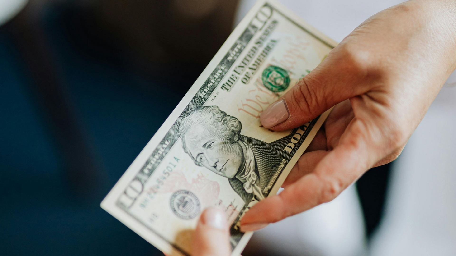 Hands holding a U.S. ten-dollar bill against a blurred blue background.
