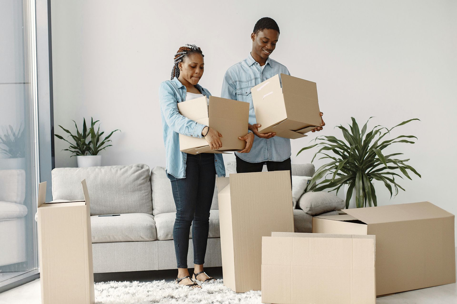Two people hold cardboard moving boxes in a bright living room furnished with a sofa and potted plants.