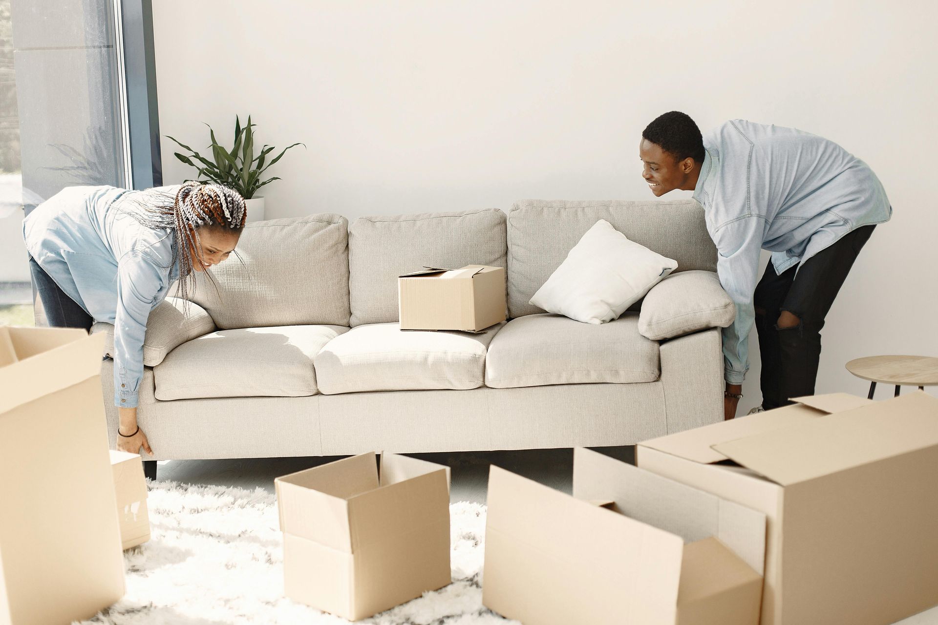 Two people lifting a beige sofa in a room filled with cardboard moving boxes.