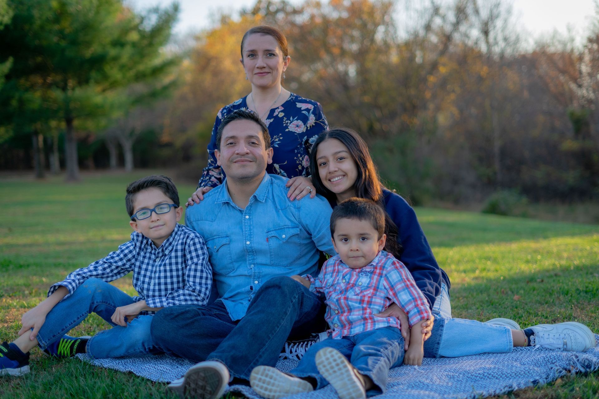 Family seated on a blanket in a sunny park, smiling at the camera.