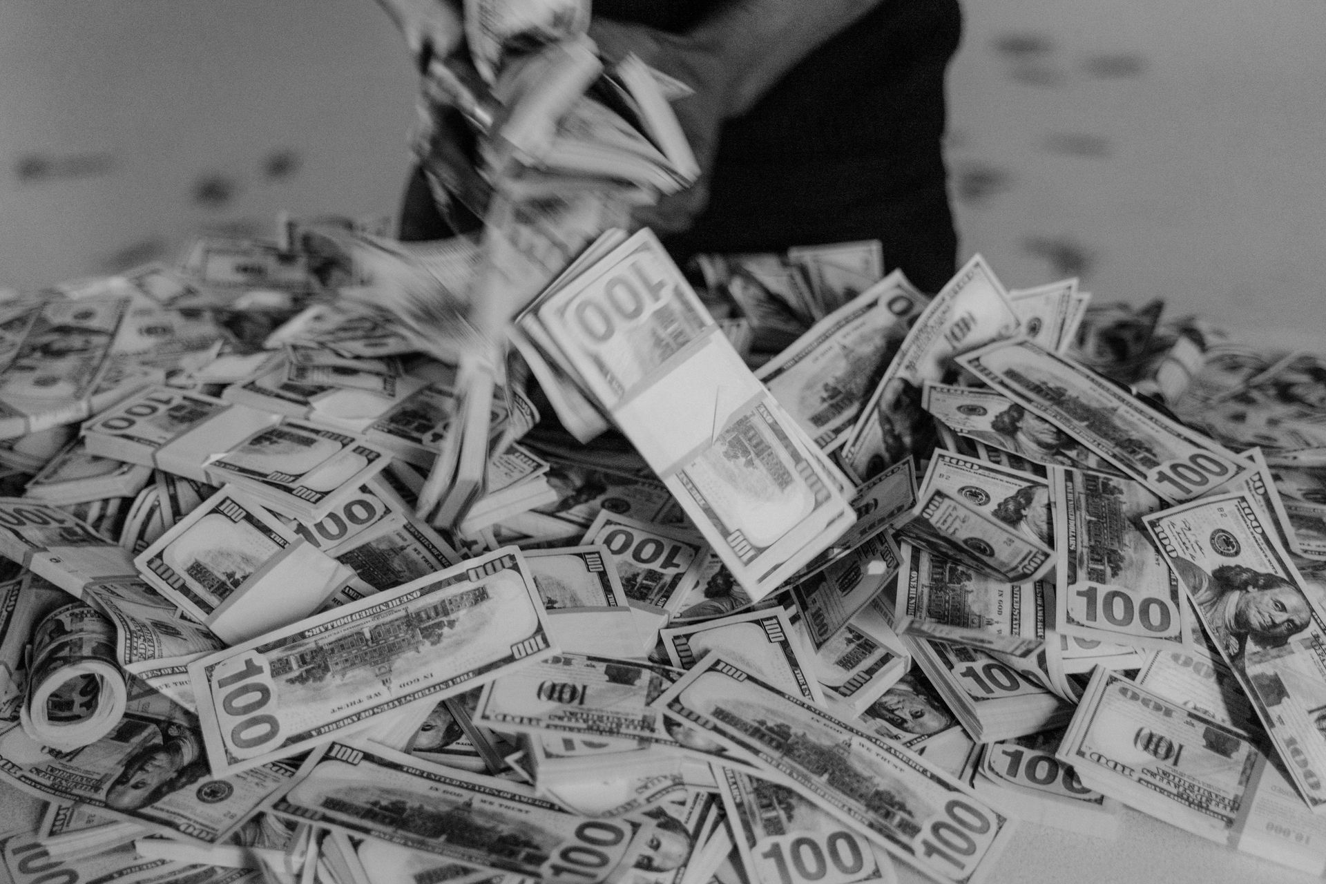 A person’s hands holding a stack of U.S. 100-dollar bills over a large, messy pile of additional currency.