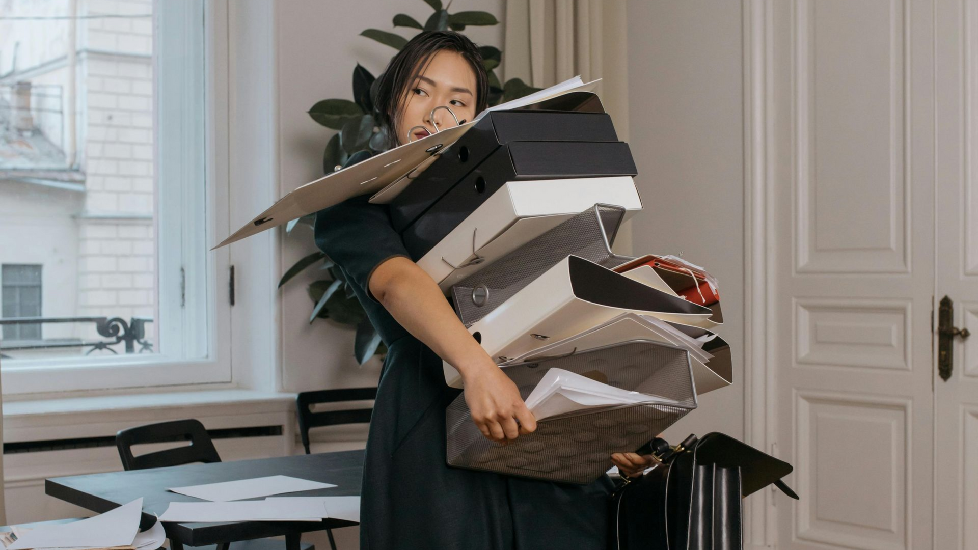 A person carries a large, unbalanced stack of office binders and papers in a bright, modern office space.