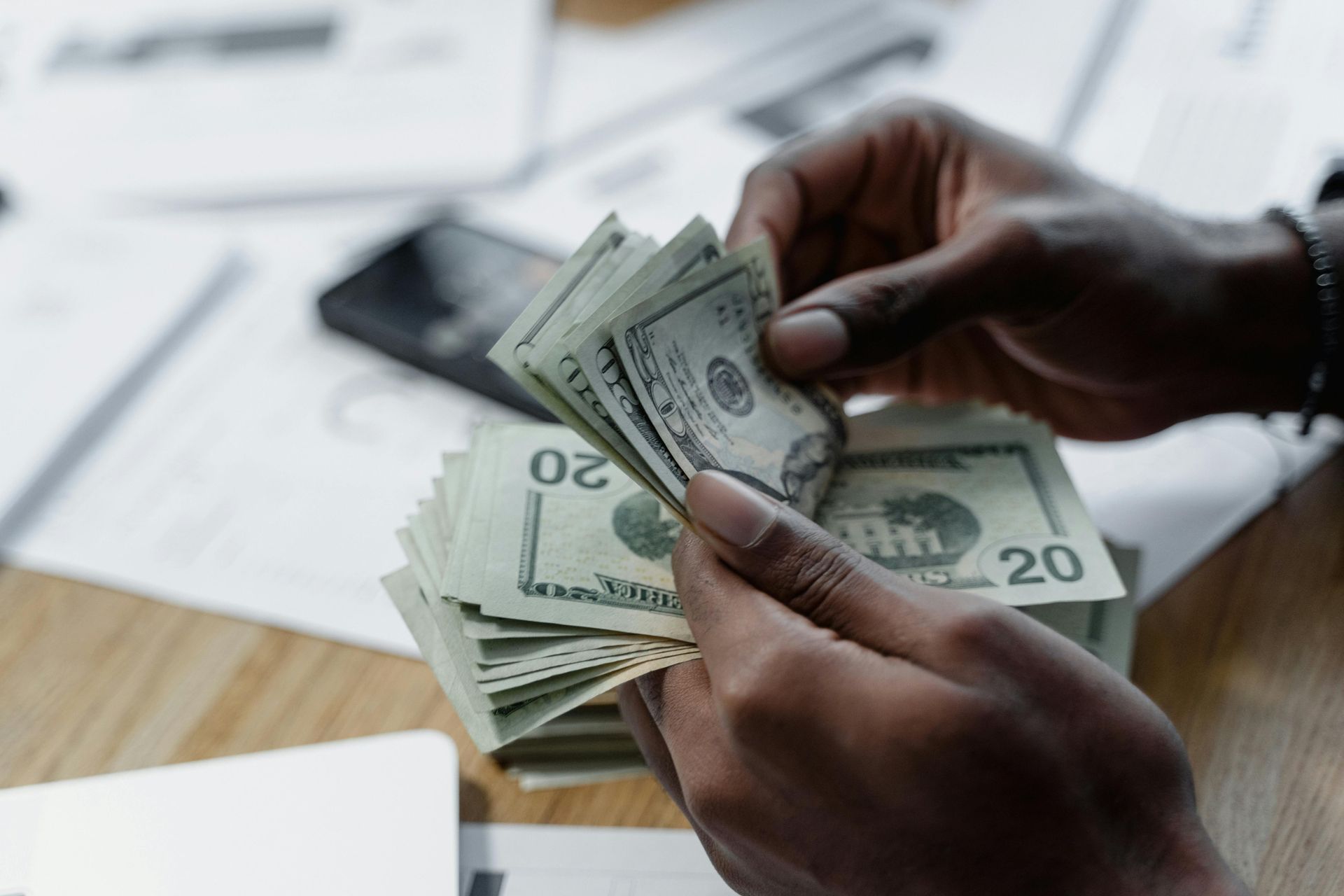 Hands counting a stack of twenty-dollar bills over a desk with documents and a calculator.
