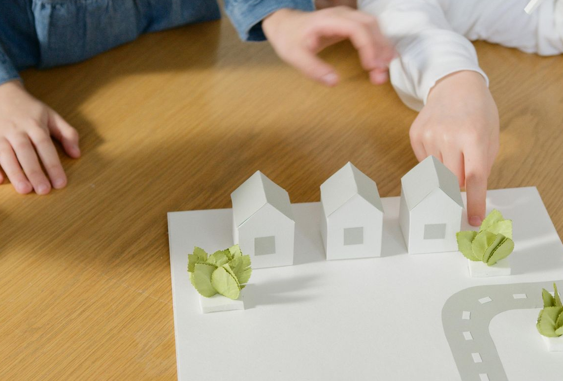 Two people building a tabletop model of a sustainable neighborhood with houses, trees, and a wind turbine.