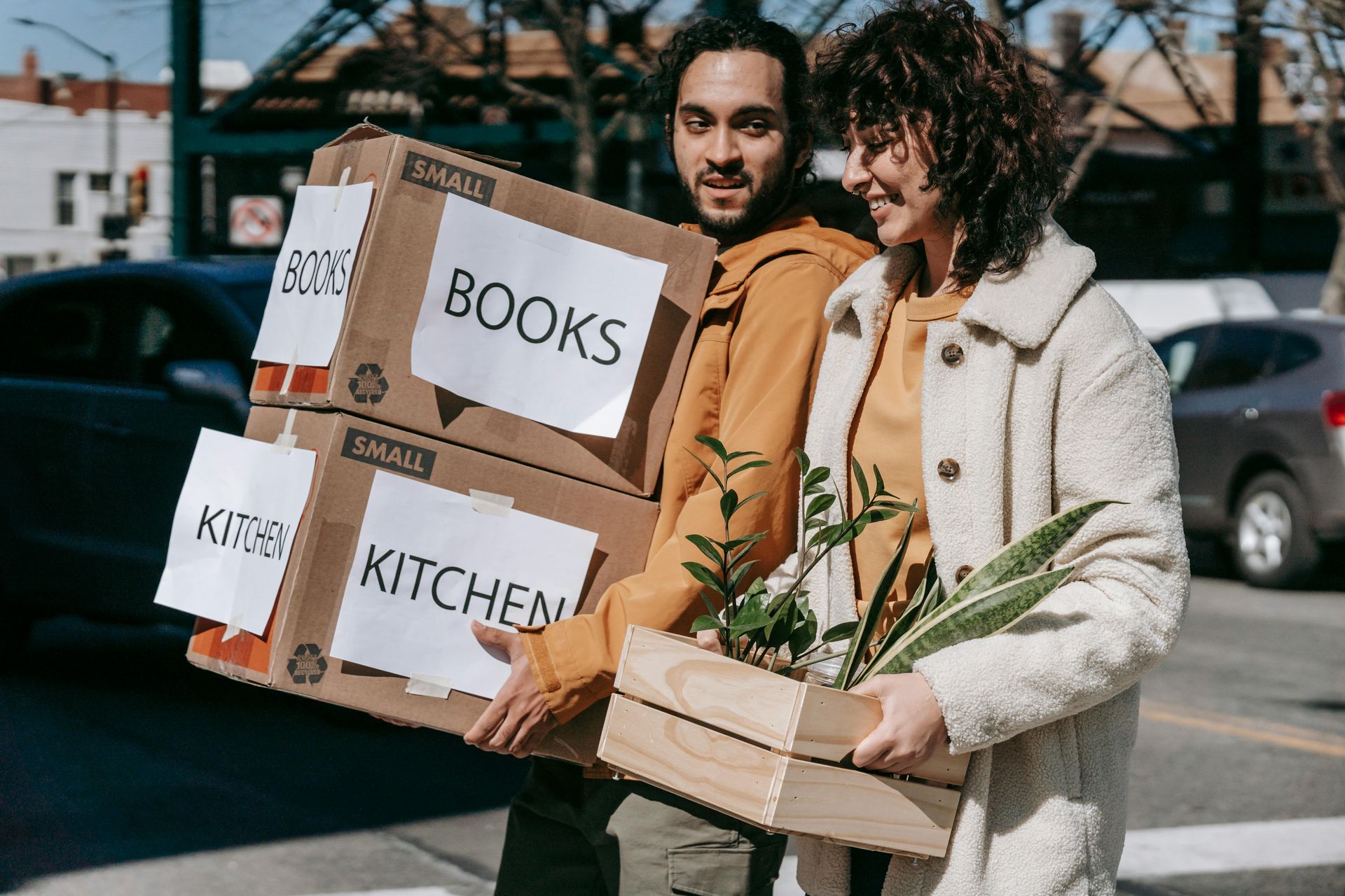 A person in a yellow hoodie and another in a white coat carry moving boxes labeled