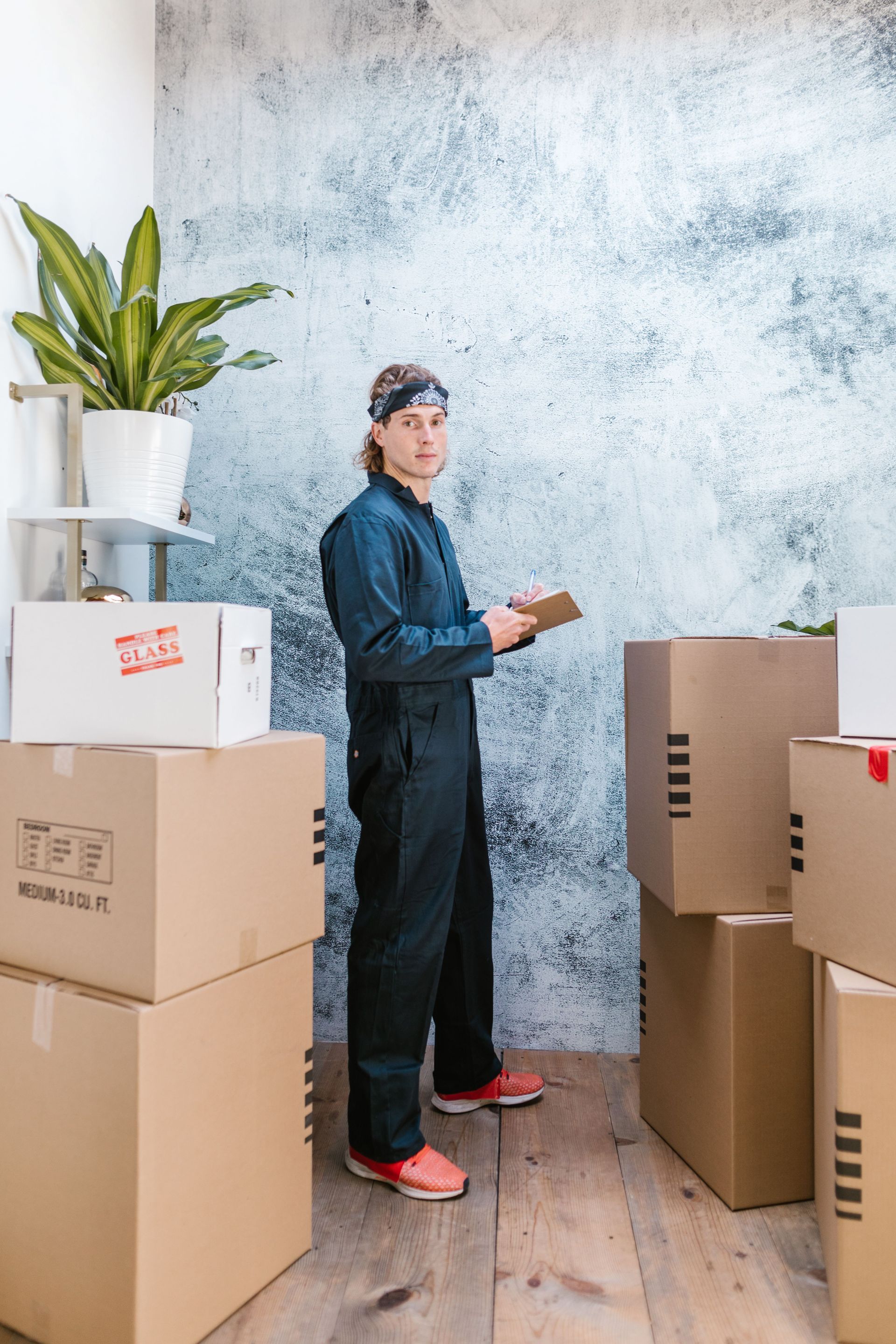 Person in dark coveralls stands among stacked moving boxes, holding a box in a storage-like room.