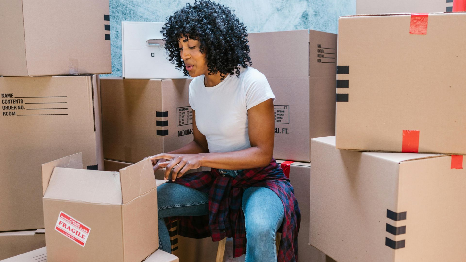 A person with curly hair wearing a white t-shirt and jeans sits among many cardboard moving boxes, taping a box shut.