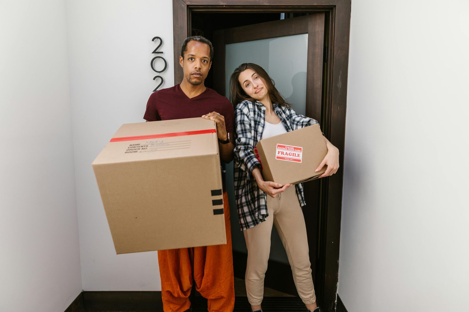 A couple stands in a doorway holding cardboard moving boxes, preparing to enter an apartment marked 202.