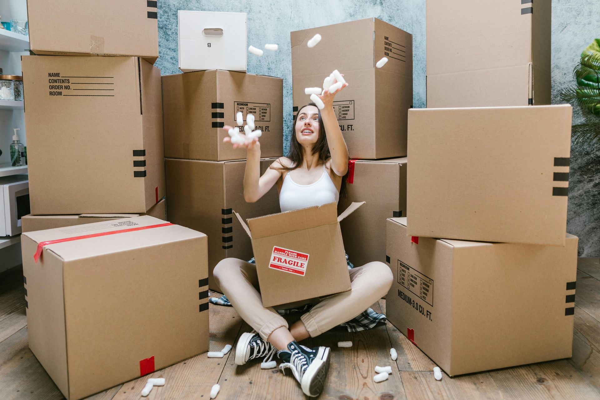 A person sitting on the floor surrounded by many cardboard boxes, joyfully tossing packing peanuts into the air.