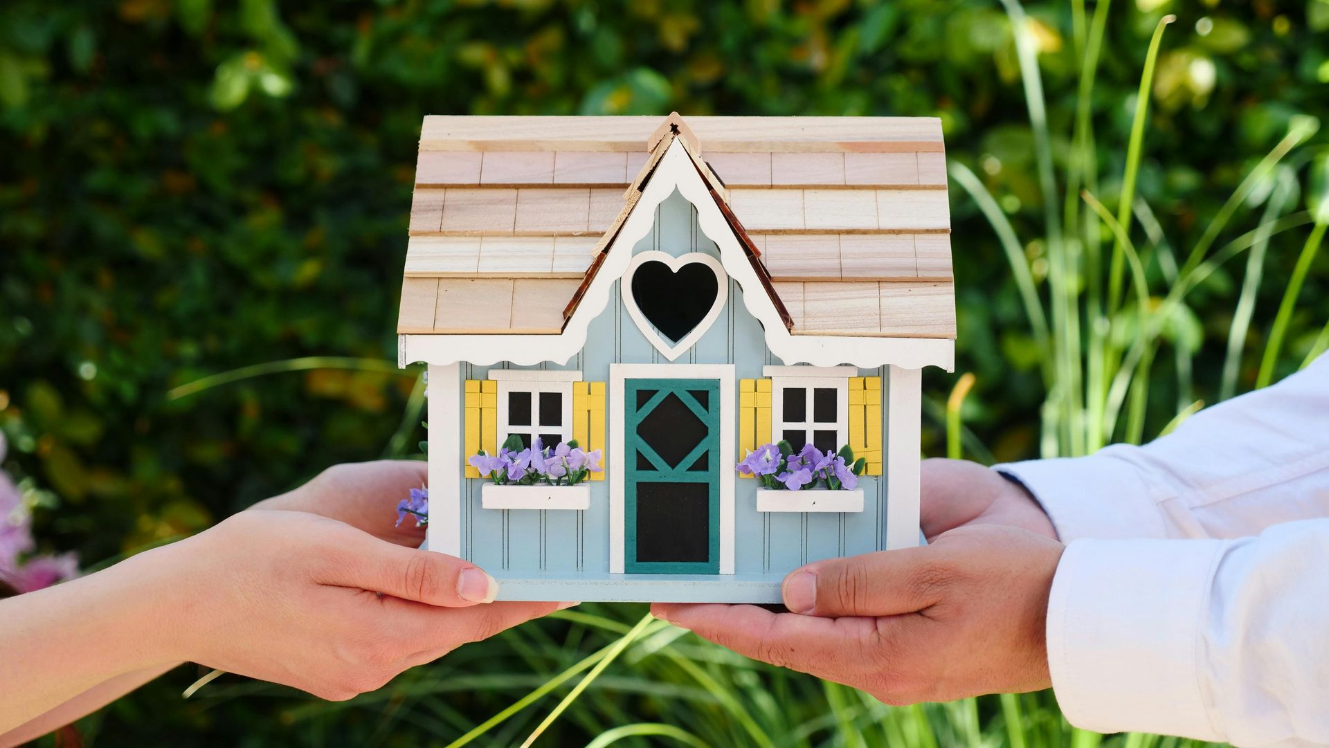 Two pairs of hands hold a small, blue decorative model house with yellow shutters against a blurred outdoor background.