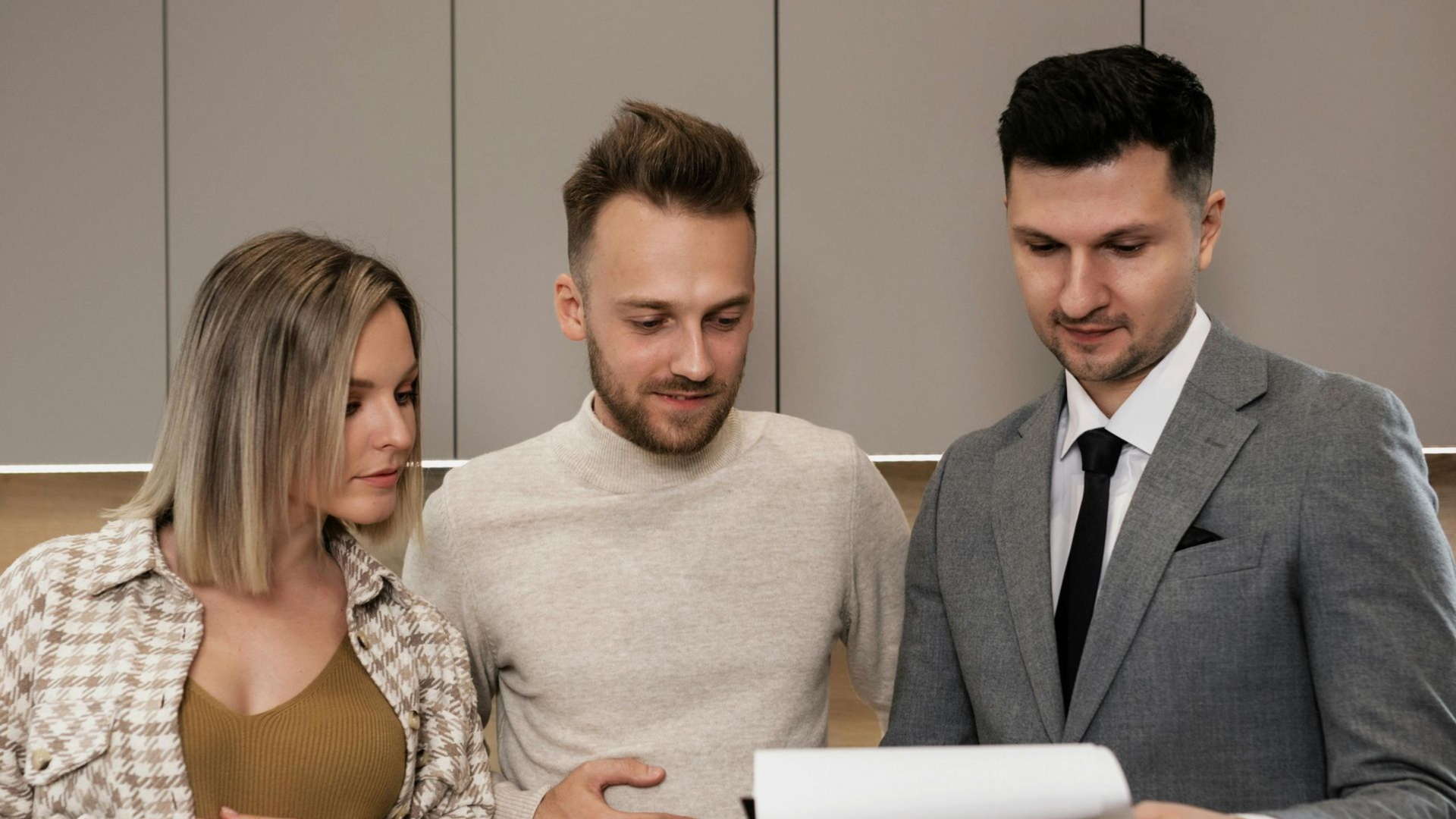 A professional in a grey suit discusses documents with two people in a modern, neutral-toned room.