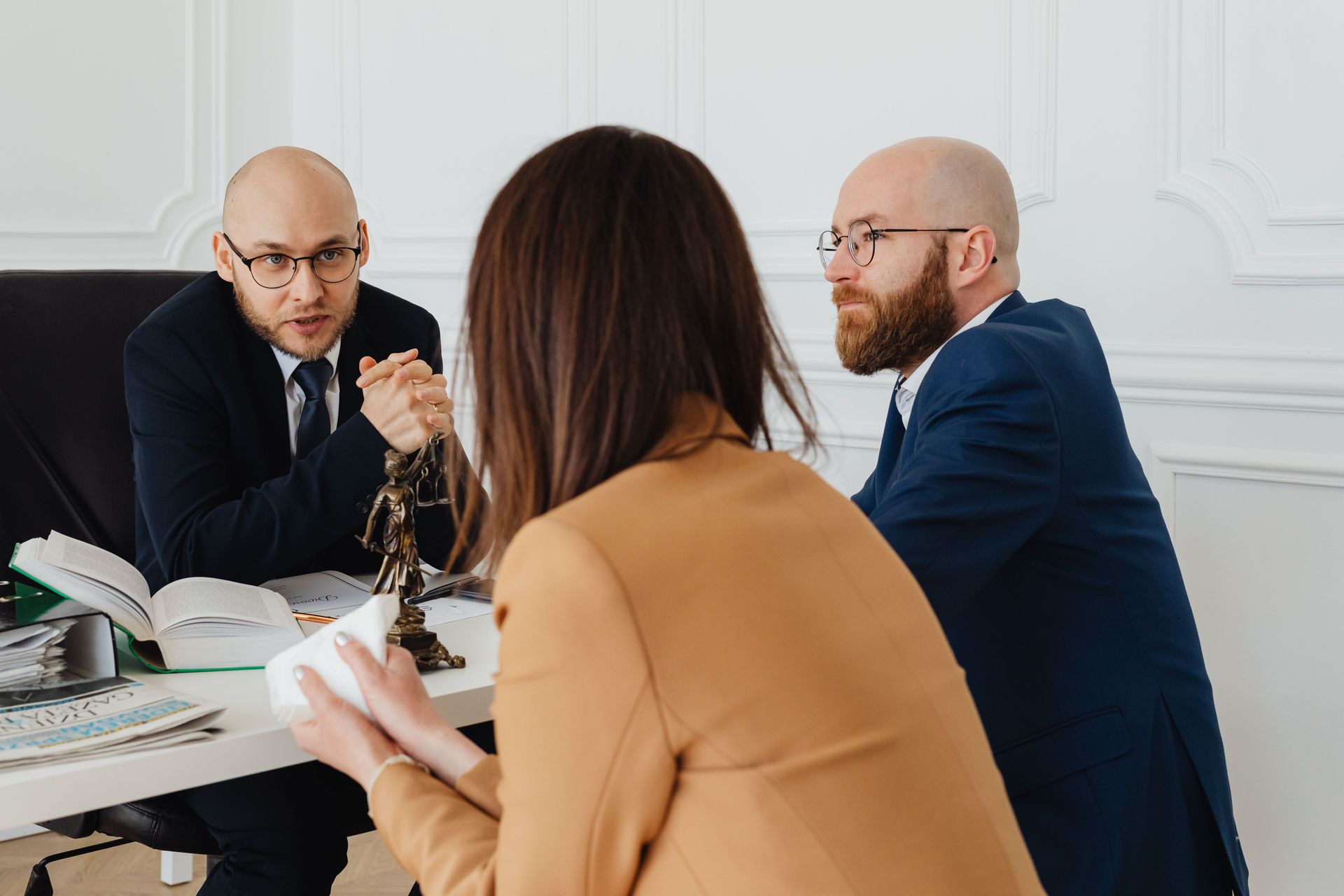 Three people in a meeting, one speaking while others listen at a table with papers and a book.